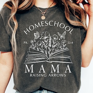 Puede incluir: Camiseta gris oscuro con las palabras "HOMESCHOOL MAMA RAISING ARROWS" en blanco. El dise&ntilde;o incluye un libro abierto con flores y mariposas. Tambi&eacute;n se incluye el texto "PR. 22:6".