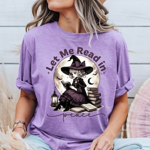 May include: A lilac-colored t-shirt featuring a graphic of a witch reading in front of a full moon. The witch is wearing a black hat and dress. The text "Let Me Read in peace" is printed on the shirt.