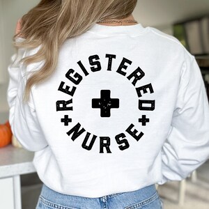 May include: White long-sleeve sweatshirt with black text that reads "Registered Nurse" in a circle with a black plus sign in the center.