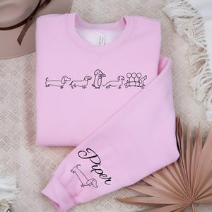 K&ouml;nnte beinhalten: Rosafarbener Pullover mit einer Reihe Dackel und dem Wort "Piper" auf dem &Auml;rmel.
