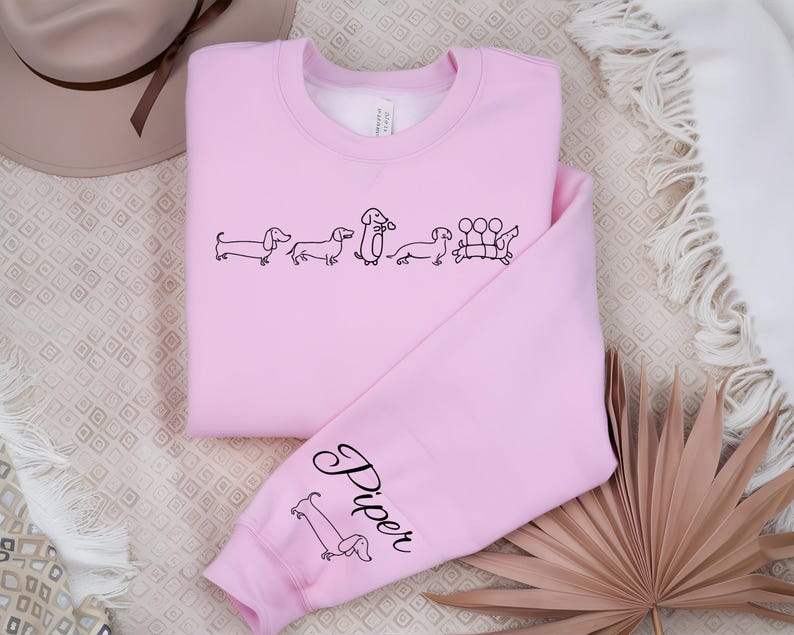 K&ouml;nnte beinhalten: Rosafarbener Pullover mit einer Reihe Dackel und dem Wort "Piper" auf dem &Auml;rmel.