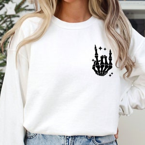 K&ouml;nnte beinhalten: Wei&szlig;er Pullover mit einer schwarzen Skeletthand, die die Rock'n'Roll-Handgeste macht. Die Hand ist von kleinen Sternen umgeben.
