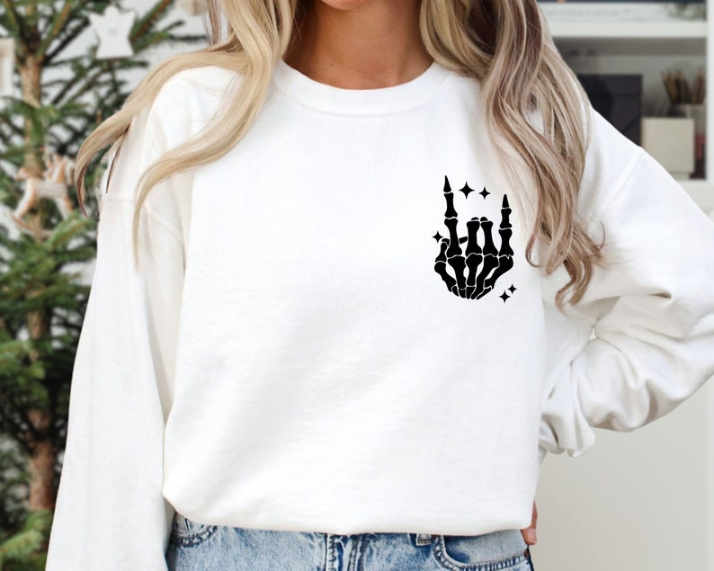 K&ouml;nnte beinhalten: Wei&szlig;er Pullover mit einer schwarzen Skeletthand, die die Rock'n'Roll-Handgeste macht. Die Hand ist von kleinen Sternen umgeben.
