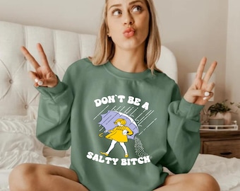 May include: Sage green sweatshirt with the text "DON'T BE A SALTY BITCH" and a graphic of a person under an umbrella, with salt pouring out. The person is wearing a yellow dress and holding a yellow umbrella.