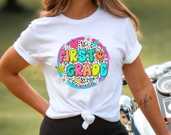 May include: White t-shirt with a colorful graphic that says "First Grade" in a bubble-like font. The graphic includes a red apple, a pencil, a paper airplane, a daisy, a lightning bolt, and a smiley face.