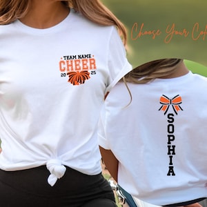 May include: A white t-shirt featuring a front graphic with the text TEAM NAME CHEER 2025 and a pom-pom icon in orange and black. The back of the shirt displays a vertical graphic with the name SOPHIA in black letters below an orange and black bow.