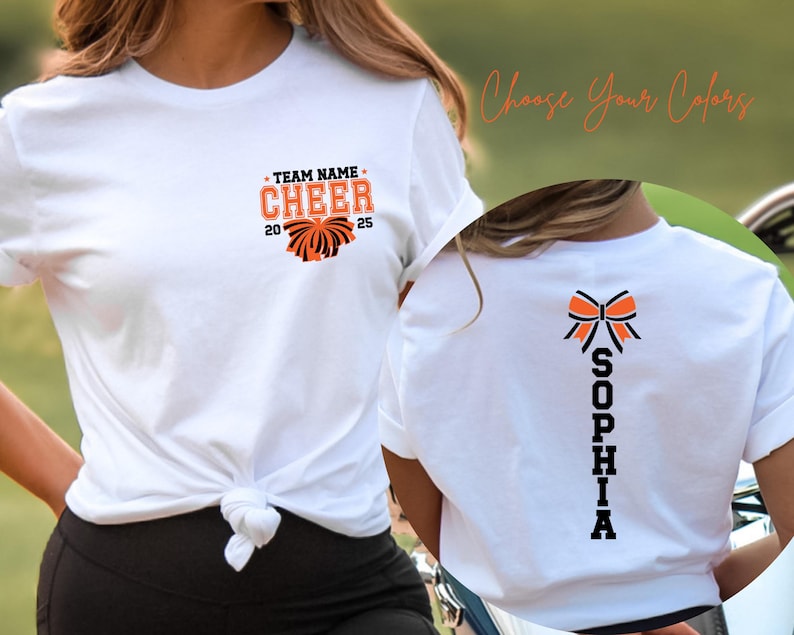 May include: A white t-shirt featuring a front graphic with the text TEAM NAME CHEER 2025 and a pom-pom icon in orange and black. The back of the shirt displays a vertical graphic with the name SOPHIA in black letters below an orange and black bow.