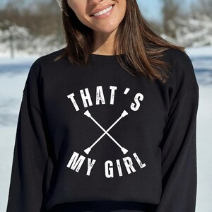 May include: Black sweatshirt with the text "THAT'S MY GIRL" in white, along with a crossed baton design. The sweatshirt is a crew neck style and is worn by a person in a snowy outdoor setting.