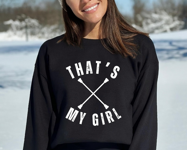 May include: Black sweatshirt with the text "THAT'S MY GIRL" in white, along with a crossed baton design. The sweatshirt is a crew neck style and is worn by a person in a snowy outdoor setting.