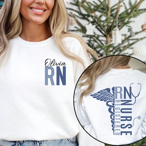 May include: White long-sleeve sweatshirt with the text "Olivia RN" in blue on the front. The back of the sweatshirt has a blue caduceus symbol with the text "RN Registered Nurse" inside.