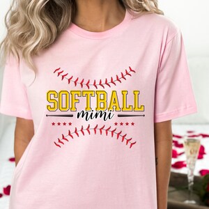 May include: Pink t-shirt with a softball graphic and the text "Softball mimi" in yellow letters.