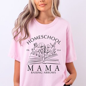 Puede incluir: Una camiseta rosa claro con las palabras "HOMESCHOOL MAMA RAISING ARROWS" en negro, junto con un dise&ntilde;o floral y de mariposas. La camiseta tambi&eacute;n incluye el texto "PR. 22:6".