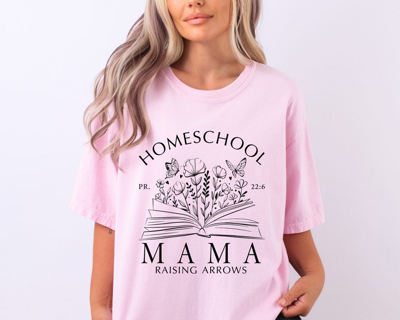 Puede incluir: Una camiseta rosa claro con las palabras "HOMESCHOOL MAMA RAISING ARROWS" en negro, junto con un dise&ntilde;o floral y de mariposas. La camiseta tambi&eacute;n incluye el texto "PR. 22:6".
