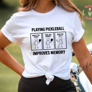May include: White t-shirt with the text "PLAYING PICKLEBALL IMPROVES MEMORY." The shirt features stick figure illustrations with the phrases "WHAT'S THE SCORE?", "WHO JUST SERVED?", and "WERE YOU 1 OR 2?" in black text.