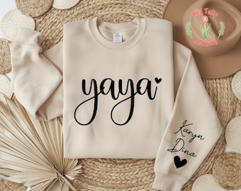 May include: A beige sweatshirt with the word "yaya" in black script and a black heart. The right sleeve has the names "Karyn Dina" in script with a black heart. The sweatshirt is a crew neck.
