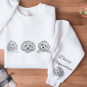 May include: White sweatshirt featuring three black and white dog portraits across the chest. The right sleeve displays the name "Daisy Clementine" in script, along with a small dog portrait. The brand logo is in the upper right corner.