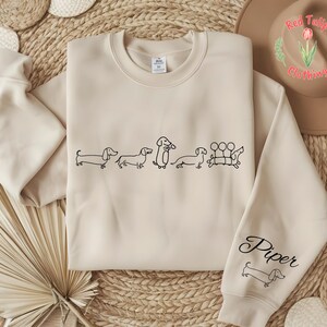 K&ouml;nnte beinhalten: Ein beiger Pullover mit einer schwarzen Umrandung einer Gruppe Dackel. Der Text "Piper" befindet sich auf dem &Auml;rmel des Pullovers.