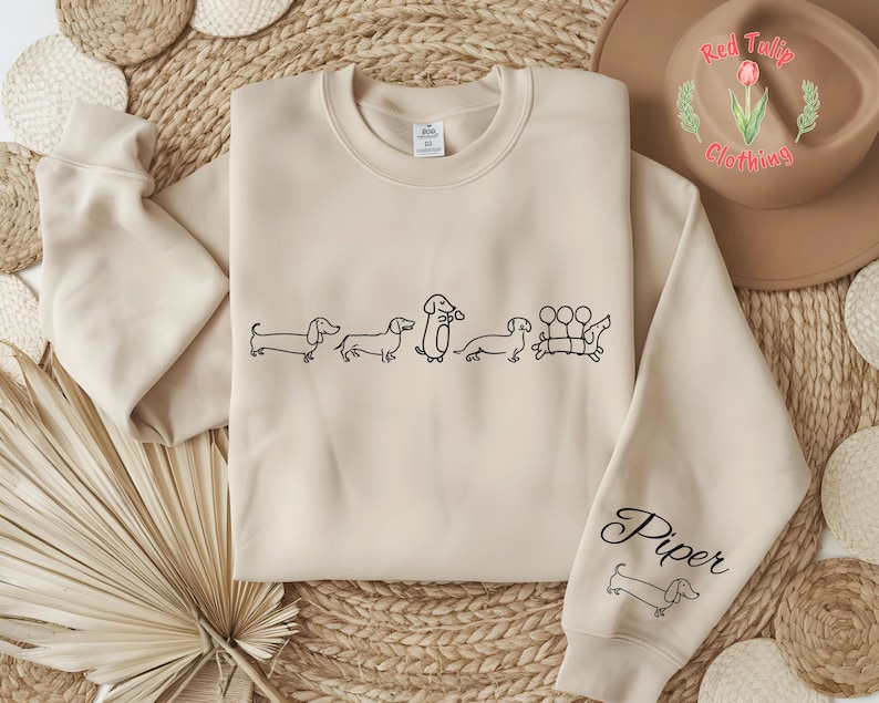 K&ouml;nnte beinhalten: Ein beiger Pullover mit einer schwarzen Umrandung einer Gruppe Dackel. Der Text "Piper" befindet sich auf dem &Auml;rmel des Pullovers.