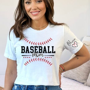 Peut inclure: T-shirt blanc avec un graphique de baseball et le texte "Baseball Mom" avec des coutures rouges. La manche porte le nom "Olivia" &eacute;crit en cursive avec un c&oelig;ur de baseball rouge et blanc.
