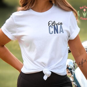 以下が含まれることがあります： 黒い筆記体で「Olivia」、青いブロック体で「CNA」と書かれた白いTシャツ。 シャツはウエストで結ばれています。 Red Tulip Clothingのロゴが右上隅に見えます。