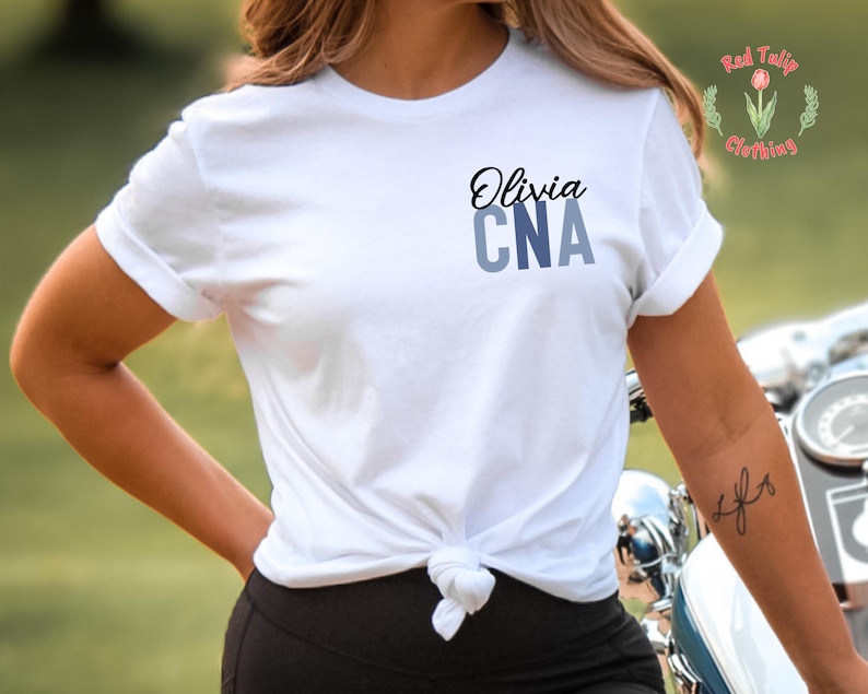 以下が含まれることがあります： 黒い筆記体で「Olivia」、青いブロック体で「CNA」と書かれた白いTシャツ。 シャツはウエストで結ばれています。 Red Tulip Clothingのロゴが右上隅に見えます。