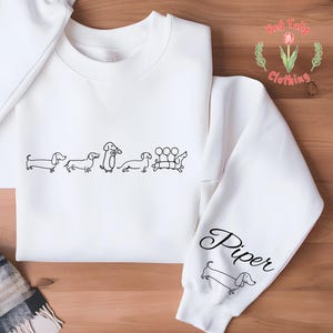 K&ouml;nnte beinhalten: Wei&szlig;er Pullover mit einer Reihe von Dackeln, die auf der Vorderseite aufgedruckt sind, und dem Namen "Piper", der auf dem &Auml;rmel aufgedruckt ist. Der Pullover wird von Red Tulip Clothing hergestellt.