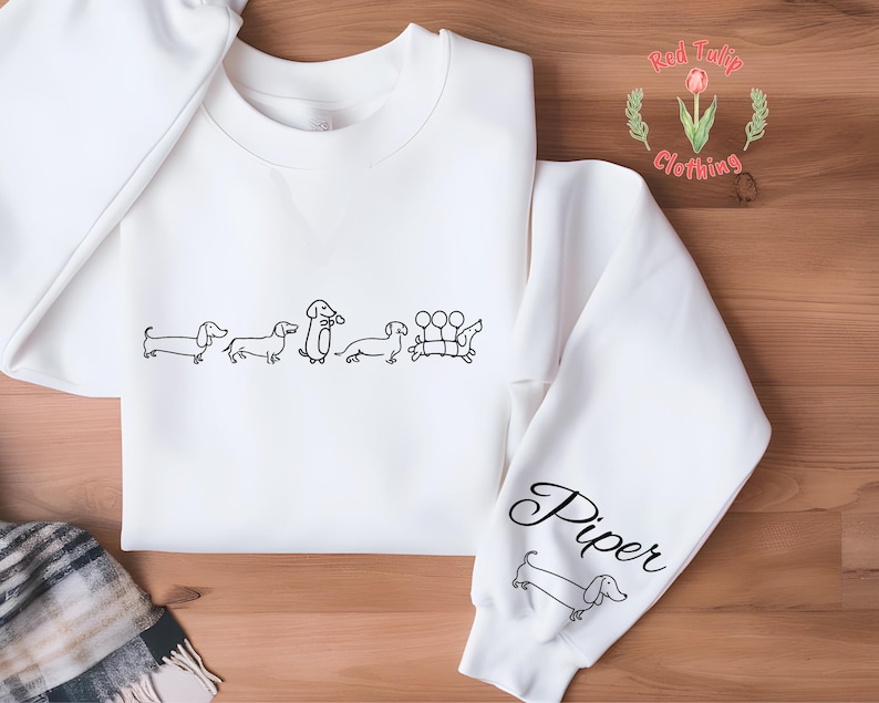 K&ouml;nnte beinhalten: Wei&szlig;er Pullover mit einer Reihe von Dackeln, die auf der Vorderseite aufgedruckt sind, und dem Namen "Piper", der auf dem &Auml;rmel aufgedruckt ist. Der Pullover wird von Red Tulip Clothing hergestellt.