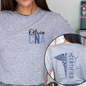以下が含まれることがあります： 前面に「Olivia」という名前と「CNA」の文字がプリントされたライトグレーのTシャツ。Tシャツの背面には、「Certified Nursing Assistant」という言葉が入った医療用カドゥケウスのデザインが施されています。