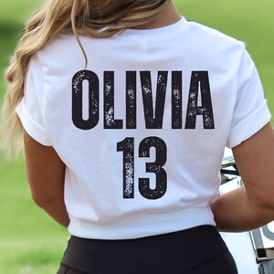 May include: White t-shirt with black text that reads "Olivia 13" on the back.