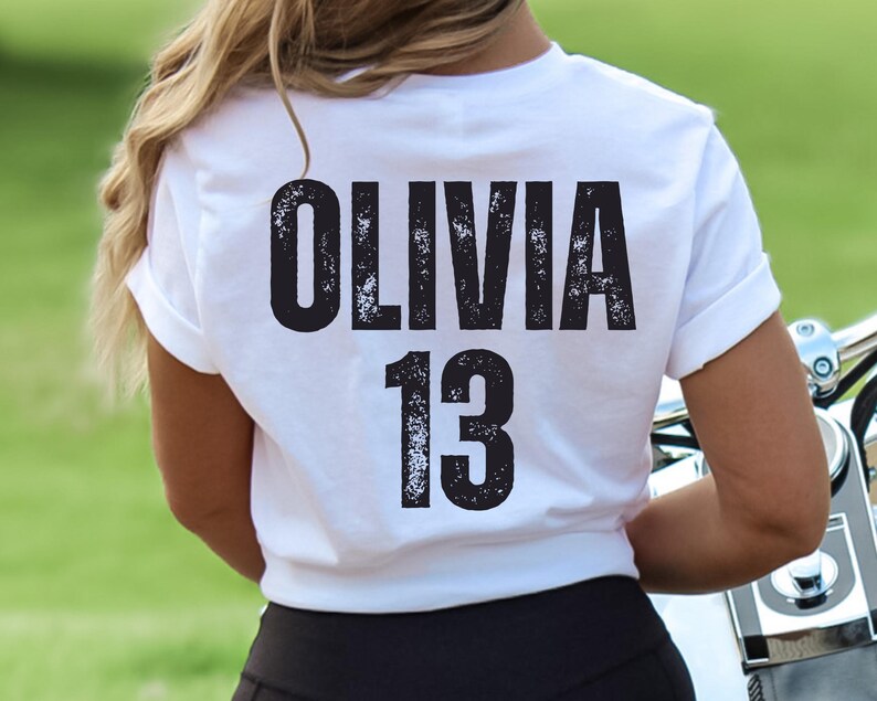 May include: White t-shirt with black text that reads "Olivia 13" on the back.