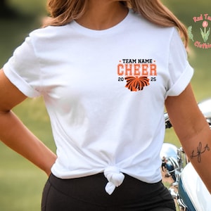 May include: A white short-sleeved t-shirt with a knotted hem. The chest features a graphic with the text "TEAM NAME CHEER 2025" in orange and black, accompanied by an orange and black pom-pom illustration. A small logo with a tulip is visible.