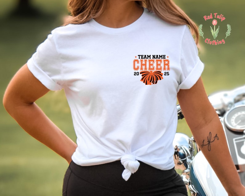 May include: A white short-sleeved t-shirt with a knotted hem. The chest features a graphic with the text "TEAM NAME CHEER 2025" in orange and black, accompanied by an orange and black pom-pom illustration. A small logo with a tulip is visible.