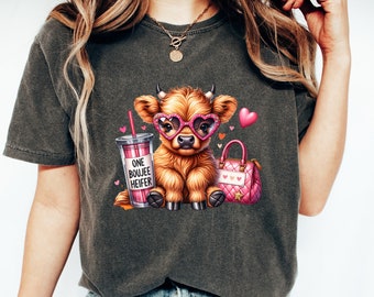 May include: A gray t-shirt with a cartoon illustration of a Highland cow wearing pink heart-shaped sunglasses, holding a pink purse and a pink and white cup with the text "One Boujee Heifer".