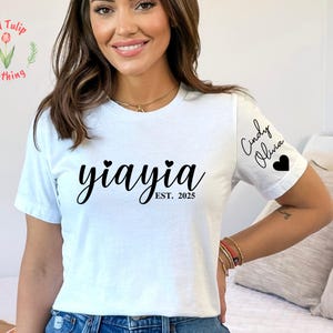 Könnte beinhalten: Weißes T-Shirt mit dem Wort "yiayia" in Schreibschrift, zwei Herzen und "EST. 2025" darunter. Der Ärmel hat die Namen "Cindy Olivia" und ein Herz. Das Shirt ist von Red Tulip Clothing.