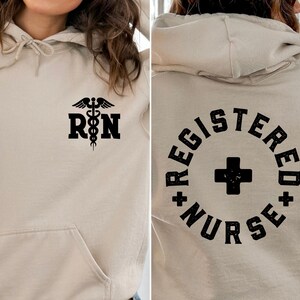 May include: A beige hoodie with a black medical symbol and the letters "RN" on the front. The back of the hoodie has a black circle with a plus sign and the text "REGISTERED NURSE".
