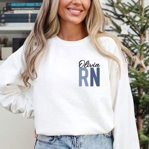 May include: White sweatshirt with the name Olivia and the letters RN in blue and gray on the front.
