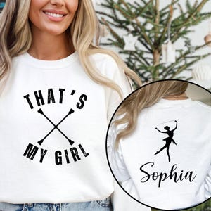 May include: White sweatshirt with the text "THAT'S MY GIRL" and crossed batons. A circular image on the back shows a silhouette of a baton twirler and the name "Sophia". The sweatshirt is a casual style.