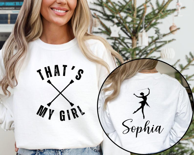 May include: White sweatshirt with the text "THAT'S MY GIRL" and crossed batons. A circular image on the back shows a silhouette of a baton twirler and the name "Sophia". The sweatshirt is a casual style.