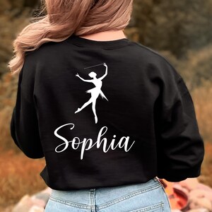May include: Black sweatshirt featuring a white silhouette of a baton twirler and the name "Sophia" in a cursive font. The design is centered on the back of the sweatshirt. The model is wearing blue jeans.