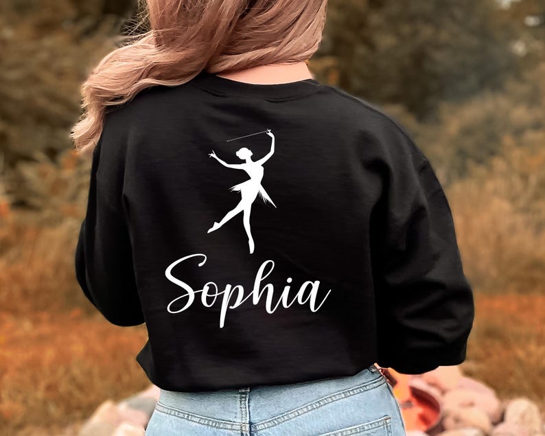 May include: Black sweatshirt featuring a white silhouette of a baton twirler and the name "Sophia" in a cursive font. The design is centered on the back of the sweatshirt. The model is wearing blue jeans.
