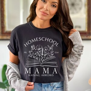 Puede incluir: Camiseta negra con la frase "HOMESCHOOL MAMA RAISING ARROWS" en blanco. El dise&ntilde;o incluye un libro abierto con ilustraciones florales y de mariposas. La camiseta es de estilo c&oacute;modo e informal.