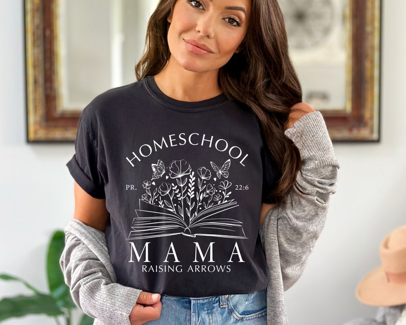 Puede incluir: Camiseta negra con la frase "HOMESCHOOL MAMA RAISING ARROWS" en blanco. El dise&ntilde;o incluye un libro abierto con ilustraciones florales y de mariposas. La camiseta es de estilo c&oacute;modo e informal.