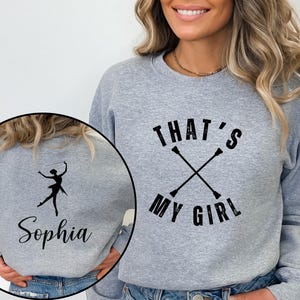 May include: A gray sweatshirt with the text "THAT'S MY GIRL" above crossed batons. A smaller image shows a silhouette of a baton twirler and the name "Sophia". The sweatshirt is a comfortable, casual style.