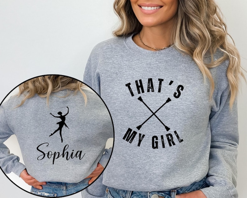 May include: A gray sweatshirt with the text "THAT'S MY GIRL" above crossed batons. A smaller image shows a silhouette of a baton twirler and the name "Sophia". The sweatshirt is a comfortable, casual style.