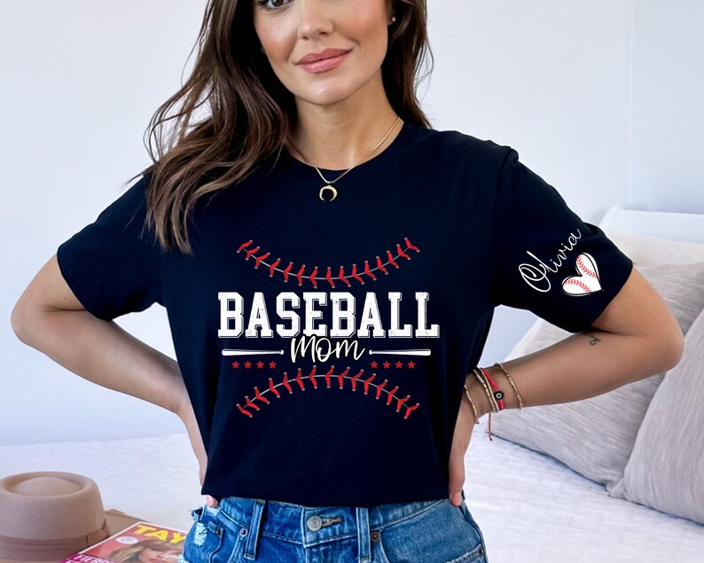 Peut inclure: T-shirt noir avec un texte blanc qui dit "Baseball Mom" et un graphique de baseball rouge cousu. Le t-shirt a un c&oelig;ur blanc avec un graphique de baseball rouge sur la manche et le nom "Olivia" &eacute;crit en cursive.