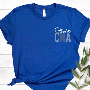 以下が含まれることがあります： ロイヤルブルーのTシャツ、袖はロールアップ。シャツには、グラデーションの青とグレーで「CNA」の文字の上に、筆記体フォントで「Olivia」という名前が書かれています。