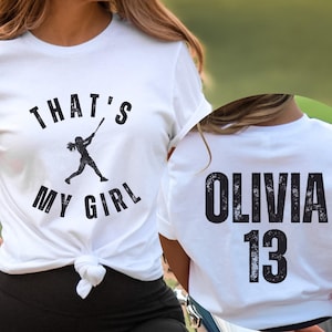 May include: White t-shirt with the text "That's My Girl" and a silhouette of a girl playing baseball. The back of the shirt has the text "Olivia 13" in a distressed font.