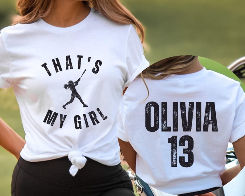 May include: White t-shirt with the text "That's My Girl" and a silhouette of a girl playing baseball. The back of the shirt has the text "Olivia 13" in a distressed font.