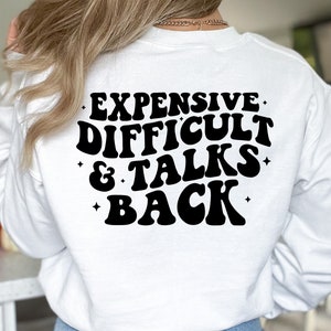K&ouml;nnte beinhalten: Wei&szlig;er Langarm-Pullover mit schwarzem Text, der "Teuer, Schwierig & Spricht Zur&uuml;ck" in einer Retro-Schriftart lautet.