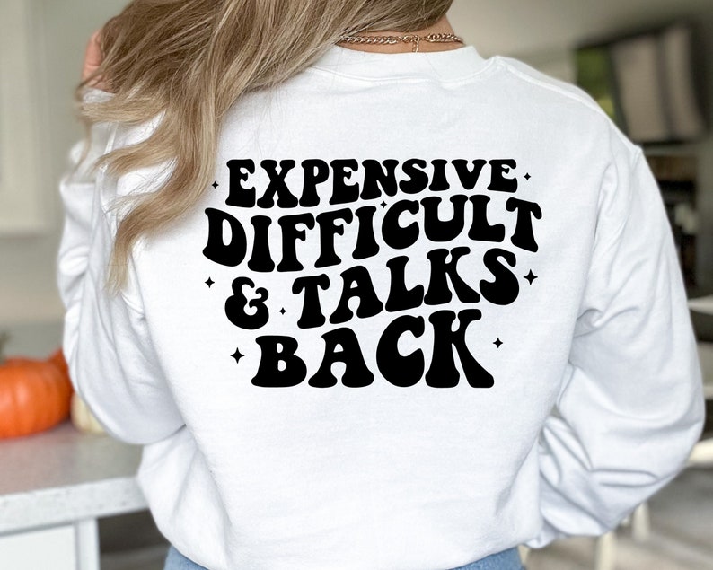 K&ouml;nnte beinhalten: Wei&szlig;er Langarm-Pullover mit schwarzem Text, der "Teuer, Schwierig & Spricht Zur&uuml;ck" in einer Retro-Schriftart lautet.